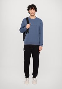 Sudadera azul de cuello redondo con texto en relieve, combinada con joggers negros tapered con bolsillos laterales y zapatillas ligeras.
