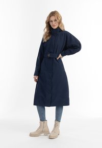 Laivaston trench coat, jossa on korkea kaulus, vyötäröltä kiristettävä vyö ja pitkät hihat; yhdistettynä vaaleansinisiin farkkuihin ja beigeihin nilkkureihin.