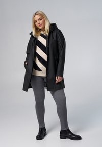 Manteau noir imperméable avec capuche, associé à un pull rayé crème et marine, un jeans gris et des bottines noires. Texture lisse.