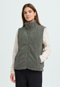 Gilet en polaire vert atténué avec col montant, fermeture éclair avant et poches latérales. Fini texturé avec une apparence douce et chaude.