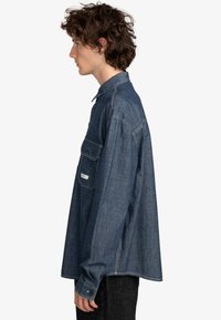 Denimskjorte i mørkeblå, med krave, lange ærmer, brystlomme og kontrasterende syninger. Bæres med sorte bukser.