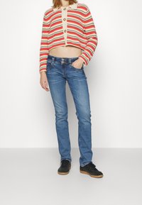 Cardigan a righe all'uncinetto in rosso e crema, con colletto e bottoni; jeans blu a vestibilità slim con chiusura a due bottoni; sneakers nere.