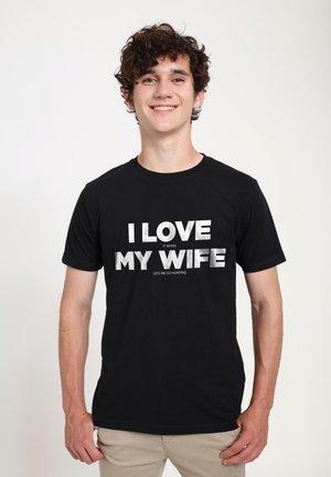Sort bomulds t-shirt med fed hvid tekst: "JEG ELSKER MIN KONE, NÅR HUN LADER MIG GÅ PÅ JAGT." Korte ærmer og rund hals.