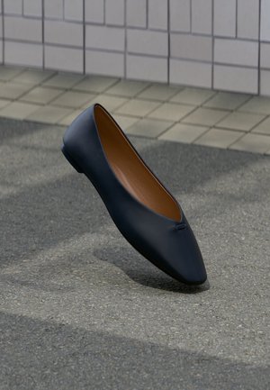 Une chaussure plate en cuir noir avec un bout pointu flottant au-dessus d'une surface asphaltée près d'un trottoir pavé.