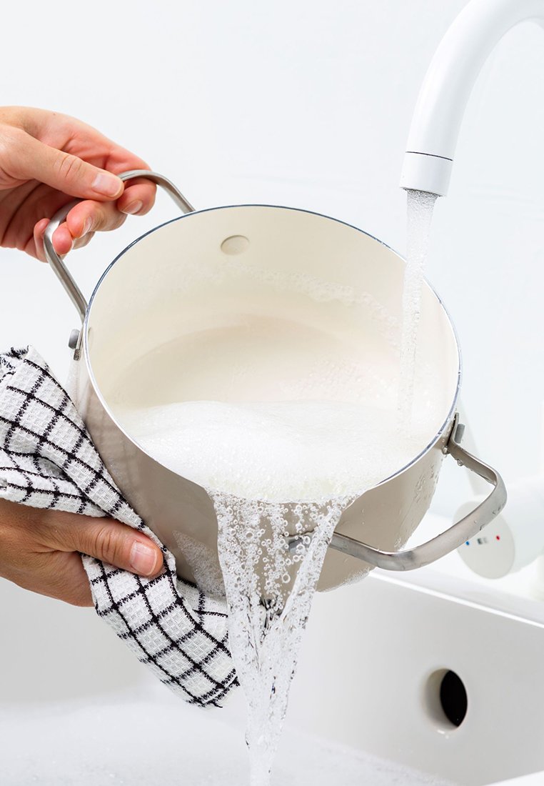 Edelstahltopf mit grauem Innenraum, gehalten von einer Hand, während Wasser aus einem weißen Wasserhahn fließt und Blasen sowie Spritzer erzeugt. Schwarz-weißer karierten Handtuch in der Hand.