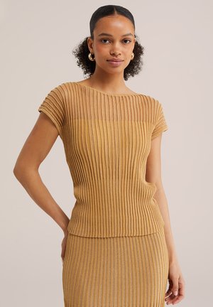 Göndör hajú nő arany karika fülbevalóval, bordázott, rövid ujjú, mustáros színű felsővel és hozzá illő szoknyával, kezét csípőn tartva áll.