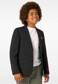 Smaidošs zēns ar cirtainiem matiem, tērpies melnā žaketē virs baltas krekla un olīvkrāsas biksēm, stāv uz balta fona.