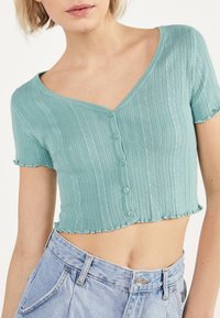 Ljus teal kortärmad cardigan med ribbad textur, V-ringning och fem knappar. Fransad nederkant ger extra detalj. Bärs med högmidjade jeans.