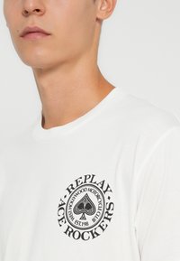 Bílý bavlněný tričko s kulatým černým grafickým motivem, na kterém je nápis "REPLAY" a "ACE ROCKERS", zobrazující symbol esa pikového ve středu.