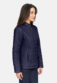 Chaqueta acolchada de color navy con cuello alto, patrones texturizados y cierre de cremallera con un acento marrón. Incluye bolsillos laterales.