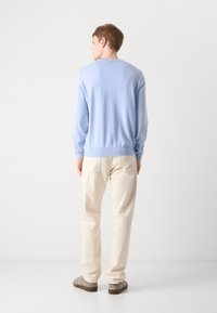 Suéter azul claro com mangas longas, punhos e bainha canelados; combinado com calças de cor creme e sapatos cinzentos, visto pela parte de trás.