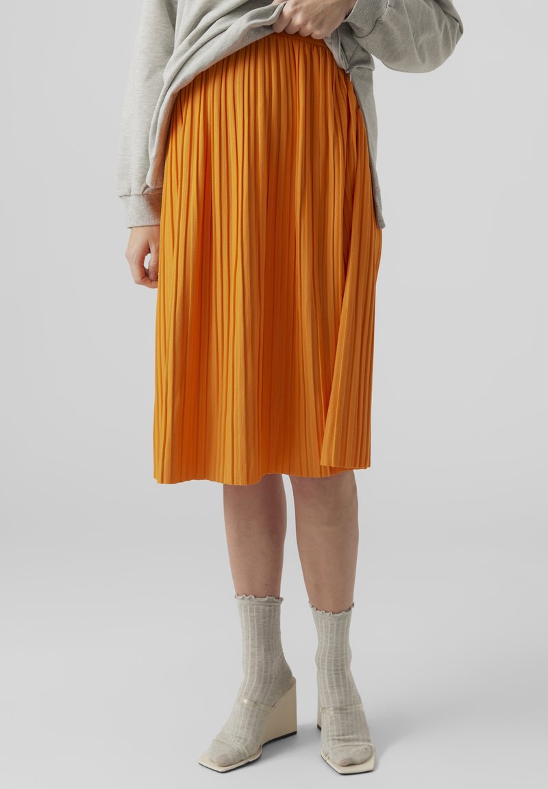 Plisserad orange midikjol med strukturerad design, kombinerad med ljusgrå ribbade strumpor och unika vita blockklackade skor.