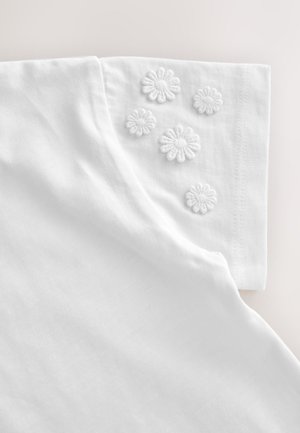 Vêtement blanc à manches courtes avec cinq petites appliques florales texturées sur la manche près de la couture de l'épaule.