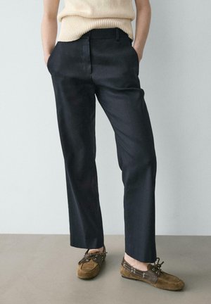 Personne portant un pantalon noir sur mesure et des mocassins en daim marron, debout les mains dans les poches sur un fond uni.