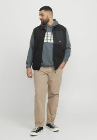 Zwarte fleecvest over een grijze hoodie met een logo, gecombineerd met beige broeken en zwarte sneakers. Casual gelaagde outfit.