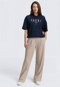 Marineblauer Kapuzenpullover mit Grafikdruck, kombiniert mit beigen Hosen mit weitem Bein und hellen Sneakers. Glatte Stofftextur.