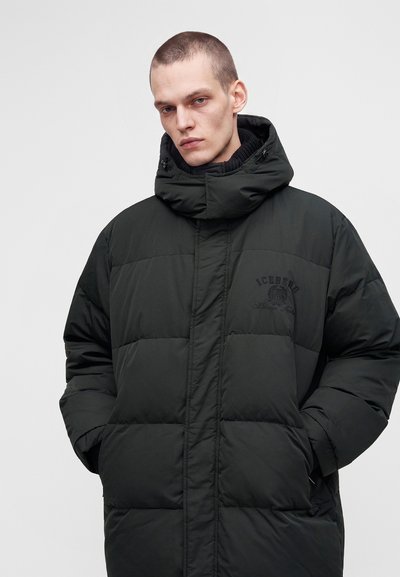 Jeune homme portant un manteau d'hiver matelassé noir avec un col montant et une capuche, mains dans les poches, fond neutre.