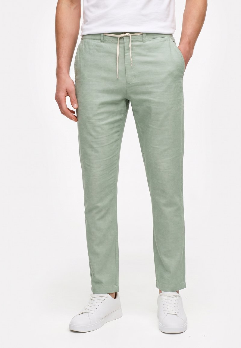 Hombre con pantalones verdes claros con cordón y bolsillos laterales, y zapatillas blancas, de pie sobre un fondo liso.