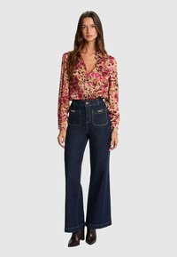 Blouse à manches longues florale dans des tons de rose et d'orange, associée à un jean flare bleu foncé avec des poches avant et des accents en métal argenté.