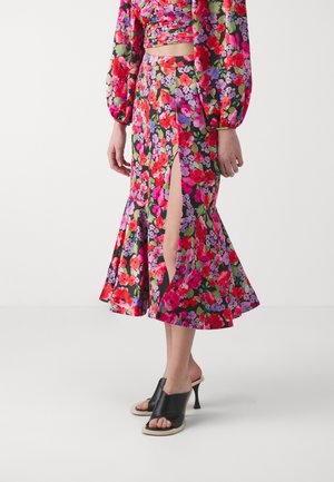 Kvetinová sukňa v živých červených, ružových a fialových odtieňoch, s rozšíreným spodným okrajom a dvoma rozparkami po stehná. Skombinovaná s čiernymi sandálmi na podpätku.