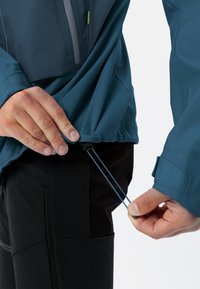 Blauwe lichte jas met een gedeeltelijke rits aan de voorkant, verstelbare zoom en elastische manchetten; gedragen met zwarte broek, handen die de trekkoorden aanpassen.