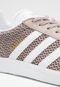 Brun vävd tygsneaker med vita ränder och en texturerad vit gummisula. Guld "GAZELLE"-logotyp på sidan, beige mockadetaljer.