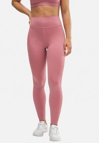 Leggings rosas de cintura alta en una tela suave y elástica, con una forma ajustada y un detalle de costura mínima, combinados con zapatillas deportivas blancas.