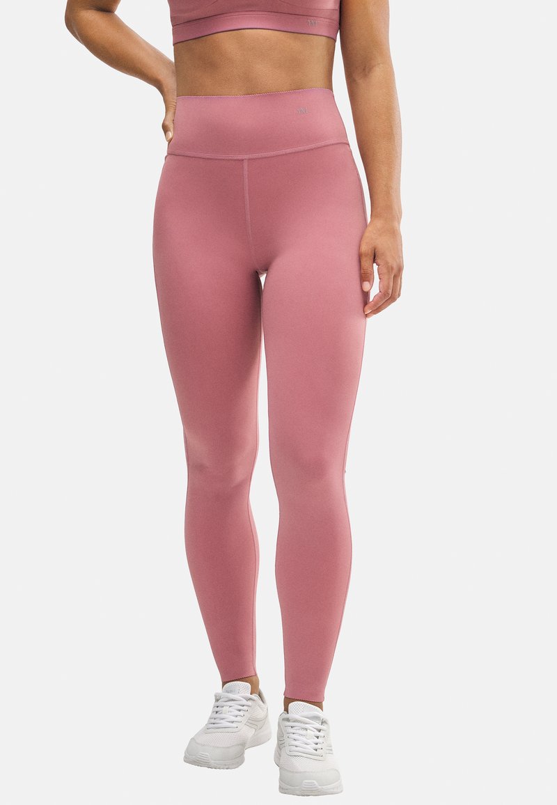 Leggings rosas de cintura alta en una tela suave y elástica, con una forma ajustada y un detalle de costura mínima, combinados con zapatillas deportivas blancas.