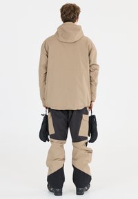 Beige vedenpitävä takki hupulla, yhdistettynä mustiin ja beigeihin lumihousuihin, joissa on suuret sivutaskut, ja käsissä pidetään lapasia.