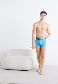 Plave bokserice s crnim pojasom na kojem se nalazi logotip "Calvin Klein". Model stoji bos kraj meke, bijele stolice na svijetlom tepihu.