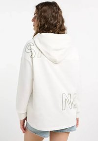 A woman with curly hair wears a white hoodie featuring large letters on the back, combined with casual denim shorts.