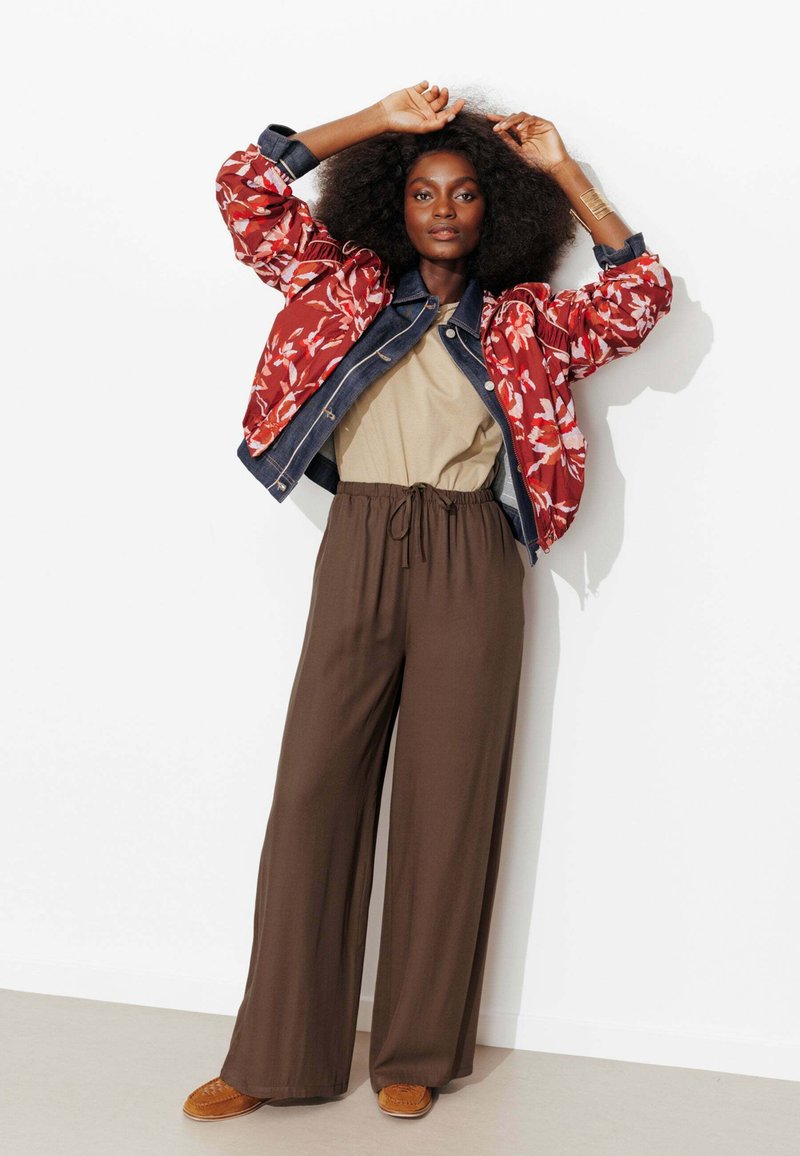 Femme aux cheveux bouclés portant une veste rouge à motifs floraux, un haut beige, un pantalon large marron et des chaussures marron, debout les bras levés contre un mur blanc.