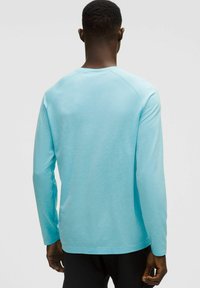 Vaaleansininen pitkähihainen paita, jossa on tekstuuripinta, pyöreä kaula-aukko ja rento istuvuus. Hihat levenevät hieman ranteista. Näytetään takanäkymä.