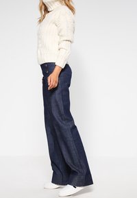 Sweter z kablem w kolorze off-white z wysokim kołnierzem, zestawiony z ciemnoniebieskimi jeansami z szerokimi nogawkami i białymi trampkami. Pojawiające się zapięcia na jeansach są widoczne.