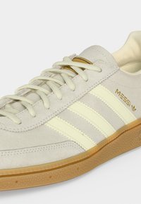 Μπεζ σουέντ αθλητικό παπούτσι με κρεμ ρίγες Adidas, λαστιχένια σόλα, λευκά κορδόνια και χρυσό λογότυπο "Messi" στο πλάι.