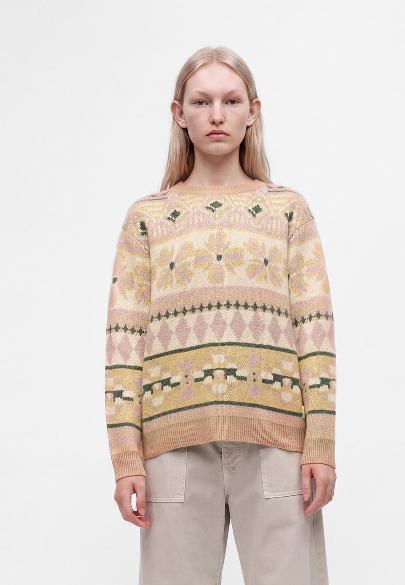 Jeune personne aux longs cheveux blonds portant un pull en maille à motif pastel et un pantalon beige clair sur un fond blanc uni.