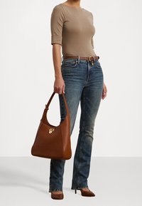 Žena v béžovém přiléhavém topu, modrých džínech s vysokým pasem, hnědém opasku a hnědých lodičkách s špičatou špičkou, drží velkou hnědou koženou kabelku.