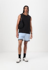 T-shirt sans manches noir, shorts en mesh bleu clair avec une bordure noire, chaussettes noires, baskets blanches avec des accents gris, et un sac en bandoulière noir.