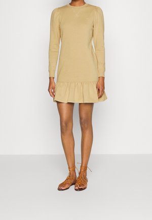 Beiges Langarmkleid mit einem Rüschensaum, aus weichem Stoff gefertigt. Das Modell trägt braune Schnürsandalen. Schlichtes, tailliertes Design.