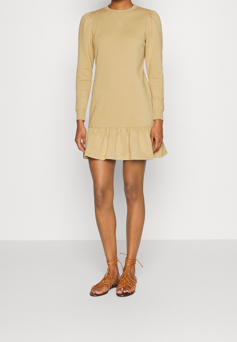 Beige šaty s dlouhým rukávem a volánkovým lemem, vyrobené z měkké látky. Model má na sobě hnědé šněrovací sandály. Jednoduchý, přiléhavý design.