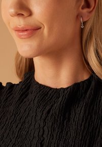 Pendientes de plata en forma de gota con un acento brillante, combinados con una blusa de tela negra texturizada. La vista en primer plano resalta el diseño de los pendientes.