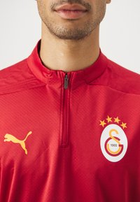 Rød Puma træningsskjorte med kvart lynlås, prydet med et gult logo og et badge med fem stjerner, designet til en tætsiddende atletisk look.