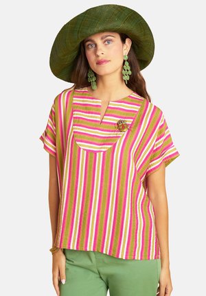 Woman wearing a green wide-brimmed hat, green leaf-shaped earrings, a pink, white, and green striped top, and light green pants.