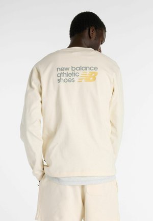 Cream long-sleeve shirt with a relaxed fit, featuring “new balance athletic shoes” printed in gray and yellow on the back.