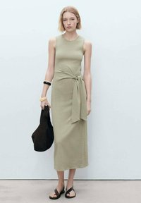 Rochie midi fără mâneci, de culoare verde măslin, cu talie legată, Fabricată dintr-un material moale. Asortată cu o geantă maro închis și sandale negre.