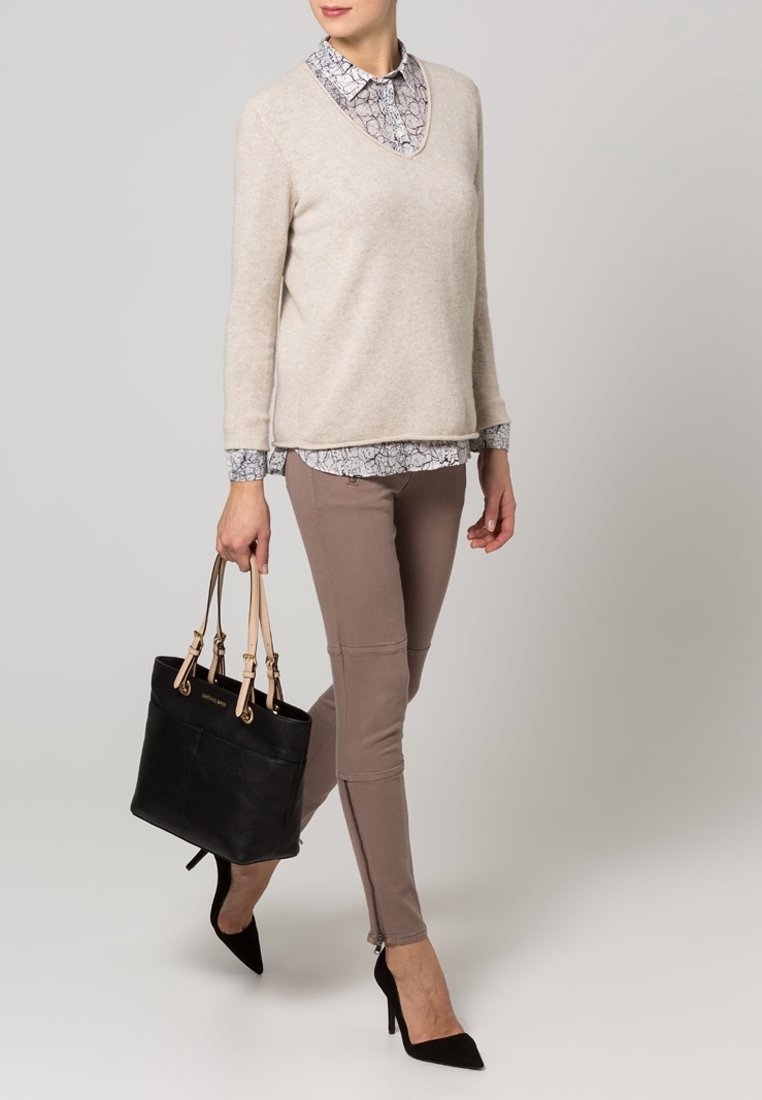 Pull beige à col en V superposé sur une chemise blanche et grise à motifs avec col, associé à un pantalon taupe ajusté et des chaussures à talons noires, tenant un sac fourre-tout noir.