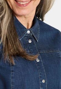 Dunkelblauses Denim-Shirt mit klassischem Kragen, ausgestattet mit silbernen Druckknöpfen und kontrastierenden Nähten, das eine glatte Textur zeigt.