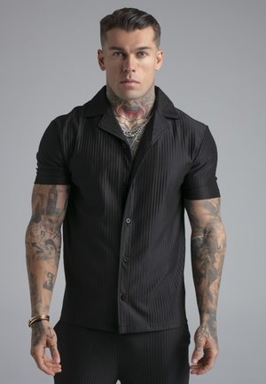 Muskuløs mand med nakke- og arm-tatoveringer iført en kortærmet sort skjorte med knapper og sølvkæder mod en ensfarvet grå baggrund.