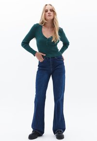 Top largo de manga larga en verde con cuello en V y botones decorativos, combinado con unos jeans de denim azules acampanados. Un calzado negro completa el conjunto.