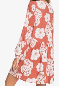 Rdzawokolorowa sukienka z przewiewnego materiału, z białymi wzorami kwiatów i liści. Długie rękawy z marszczonymi mankietami i rozkloszowanym dołem.